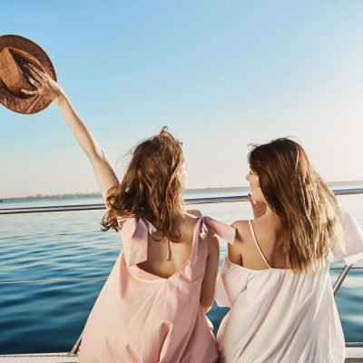 Back portrait of two female friends sitting on boat, waving with hat while talking and enjoying looking at seaside. Sisters finally took vacation to visit their mom who lives in Italy.
