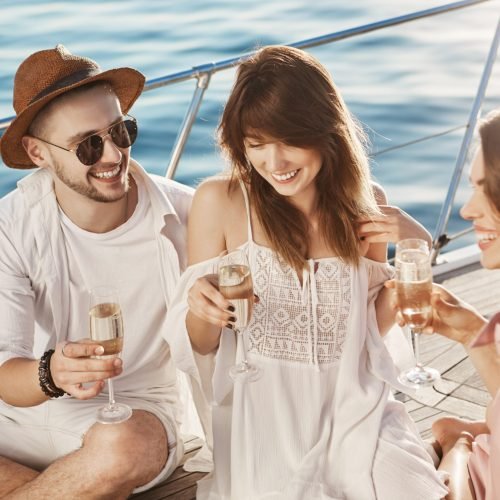 Close-up outdoor portrait of three friends talking and drinking champagne while sitting on board of boat and enjoying sunlight. Young couple and their friend discussing wedding ceremony. Vacation and emotions concept