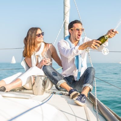 Couple celebrating anniversary with champagne on a boat - Attractive caucasian man uncorking champagne and having party with her girlfriend on vacation - Two young tourists having fun on a boat tour in the summertime
