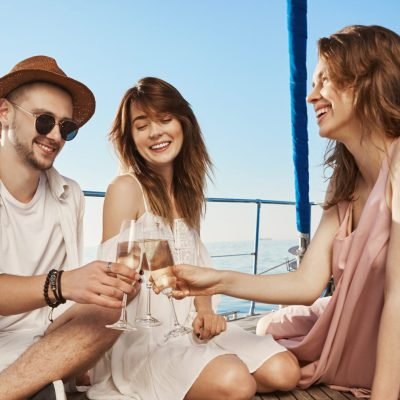 Outdoor shot of three trendy european friends sitting on boat, having lunch and drinking champagne, expressing joy and pleasure. Every year they book tickets to warm countries in winter. Copy space.